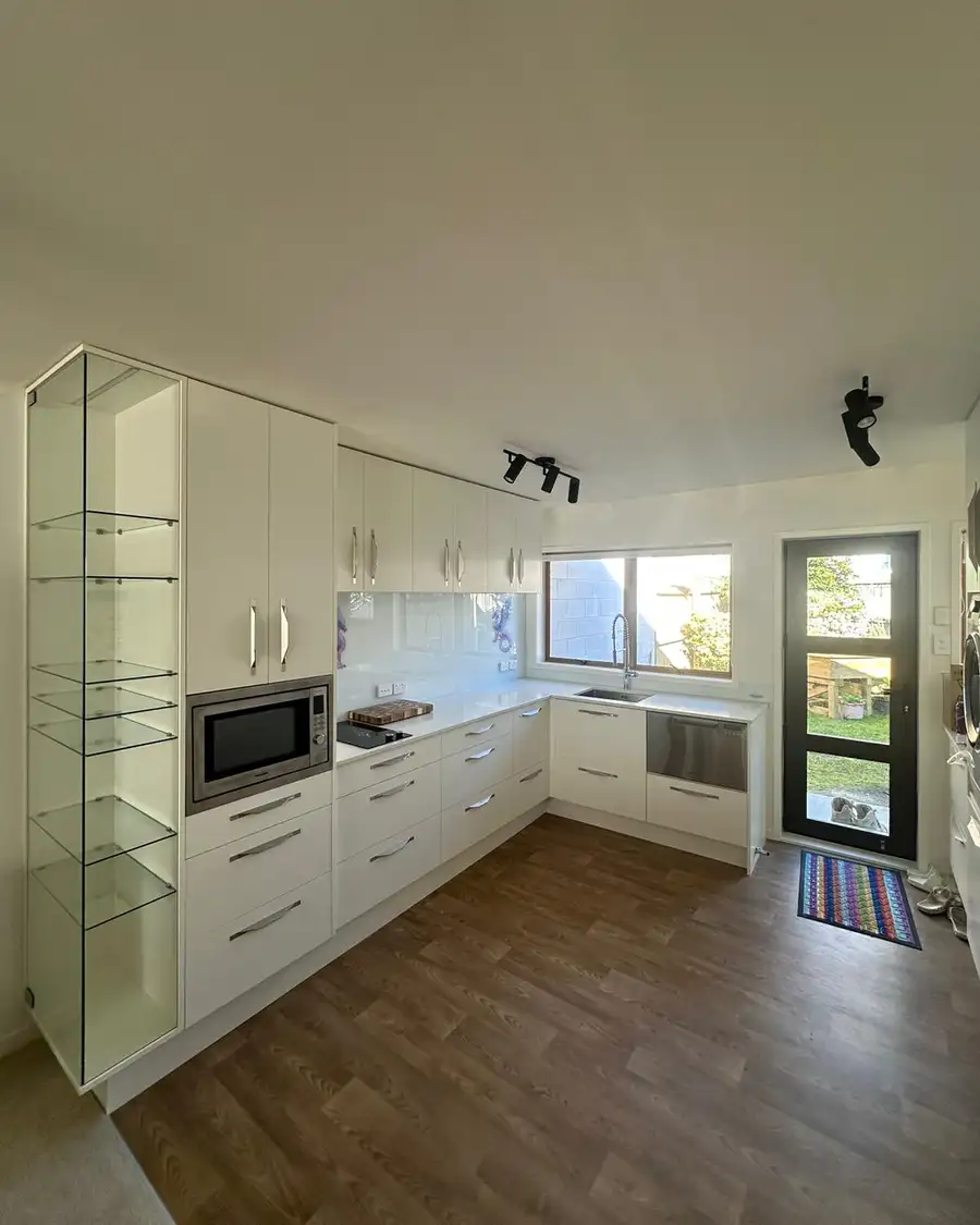 The finished Te Atatū Peninsula kitchen, white cabinets with pull handles, glass display shelves, a built-in microwave, the hob and the sink under the window.